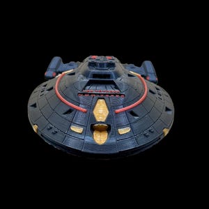以下が含まれることがあります： スタートレックのUSSヴォイジャー宇宙船の濃いグレーの模型。モデルは赤と金のアクセントがあり、船の名前が前面に見えます。船のデザインには、円形の一次船体と角張った二次船体が含まれています。