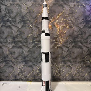May include: A detailed model of the Saturn V rocket, mainly white with black detailing and the USA emblem. The model is on a black base with the words "SATURN V" in white. The rocket design includes multiple stages and engine details.