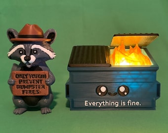 May include: A whimsical figurine set featuring a raccoon wearing a ranger hat and holding a sign that reads, "ONLY YOU CAN PREVENT DUMPSTER FIRES." Beside it is a blue dumpster with flames inside and the text "Everything is fine."