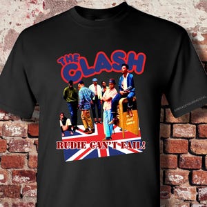 May include: Black t-shirt featuring a graphic of The Clash band members. The design includes the band's name in red and blue, and the phrase "RUDIE CAN'T FAIL!" over a Union Jack flag. The shirt is displayed against a brick wall.