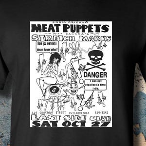 May include: A black short-sleeved t-shirt featuring a white graphic print of a concert flyer. The design includes the text Meat Puppets, Stretch Marks, East Side Club, Sat Oct 27, and various illustrations including a skull, an eight ball, and figures.