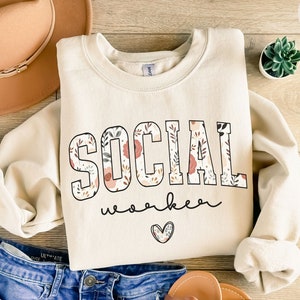 May include: A beige sweatshirt with the words "SOCIAL WORKER" printed in black with a floral pattern inside the letters. A small heart is below the text.