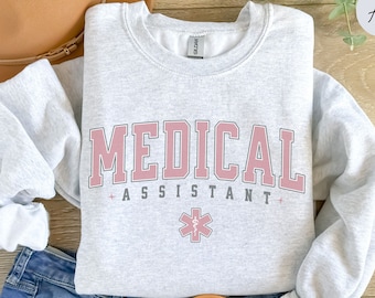 May include: Gray sweatshirt with the text "MEDICAL ASSISTANT" in pink letters with a red medical symbol.
