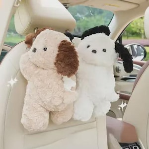 May include: Two plush dog-shaped car seat organisers. One is beige with brown ear accents, the other is white with black ear accents. Both have button eyes and are designed to hang from a car seat headrest.