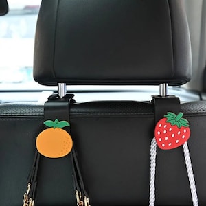 May include: Two car seat headrest hooks with fruit designs. One hook has an orange design and the other has a strawberry design. Both hooks have a silver metal hook and a black plastic base.