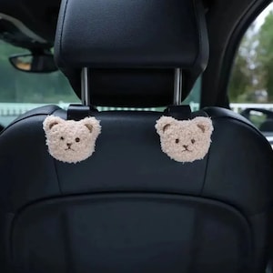 Puede incluir: Dos colgadores beige para reposacabezas de asiento de coche con forma de osito de peluche. Las caras de los osos de peluche tienen rasgos bordados y están unidas a correas negras. El asiento del coche es negro.
