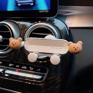 May include: White car phone holder with two brown bear-shaped clips. The holder is attached to the air vent in the car. The text "Hi I'm Little One" is printed on the holder.