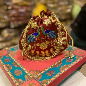 May include: A maroon drawstring bag with gold embroidery of peacocks and pearl trim. The bag is sitting on a red and blue box with a floral design.
