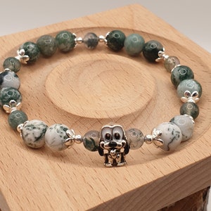Könnte beinhalten: Armband mit grün-weiß gesprenkelten Steinperlen, unterbrochen von silberfarbenen floralen Akzenten. Ein kleiner, silberfarbener Hundeanhänger befindet sich in der Mitte. Das Armband liegt auf einer Holzoberfläche.