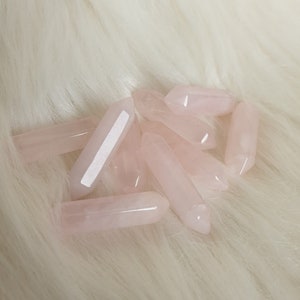 Könnte beinhalten: Eine Ansammlung hellrosa Rosenquarz-Kristallspitzen. Die Kristalle sind poliert und haben ein glattes, durchscheinendes Aussehen. Sie sind auf einer weichen, weißen, flauschigen Oberfläche angeordnet.
