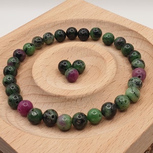 Könnte beinhalten: Eine Ansammlung runder Edelsteinperlen in Grün- und Rubinrottönen, kreisförmig angeordnet. Die Perlen werden auf einem hellen Holztablett präsentiert, mit einer herzförmigen Anordnung von Perlen in der Mitte.