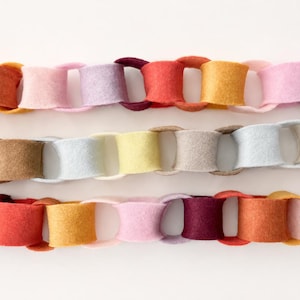 May include: Three horizontal rows of interlocking felt chain links in a variety of colours. The top row features shades of red, orange, yellow, pink, and purple. The middle row displays neutral tones of cream, tan, light blue, and yellow. The bottom row repeats the vibrant colour palette of the top row.