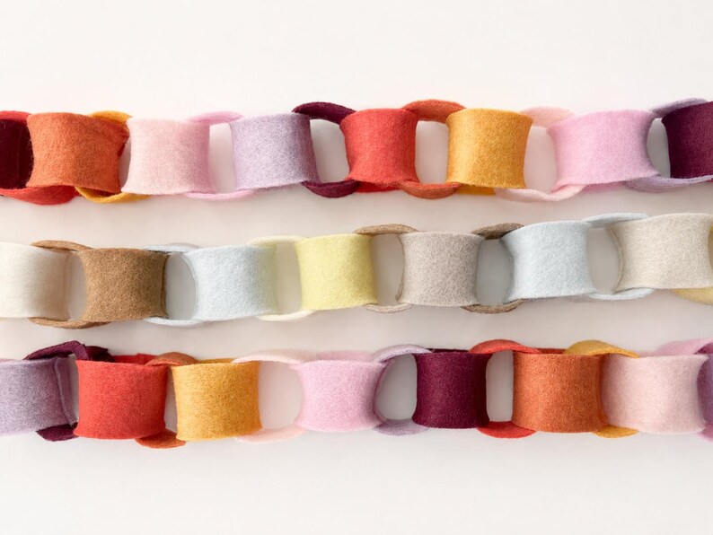 May include: Three horizontal rows of interlocking felt chain links in a variety of colours. The top row features shades of red, orange, yellow, pink, and purple. The middle row displays neutral tones of cream, tan, light blue, and yellow. The bottom row repeats the vibrant colour palette of the top row.