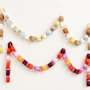 May include: Two decorative paper chains arranged in a draped formation. The top chain features muted tones of beige, light blue, white, yellow, and tan. The bottom chain consists of vibrant colours including red, orange, pink, purple, and magenta.