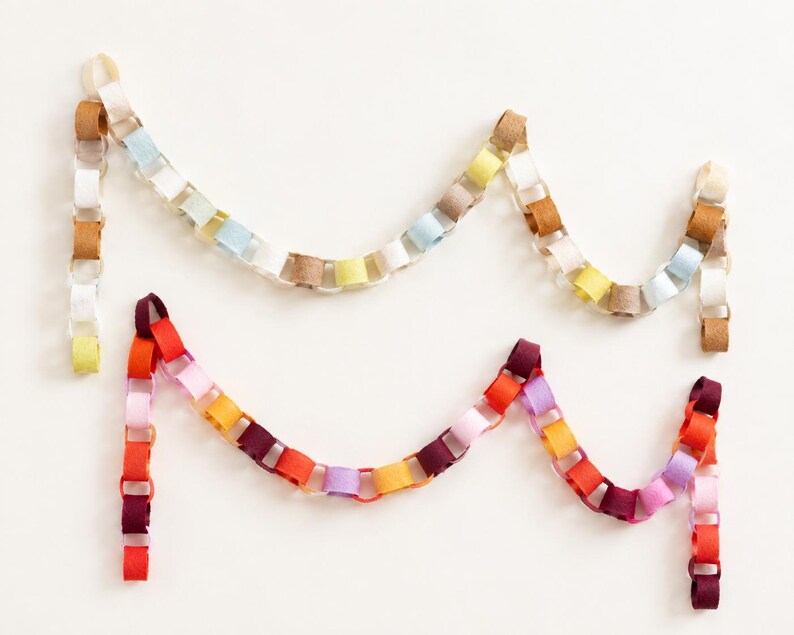 May include: Two decorative paper chains arranged in a draped formation. The top chain features muted tones of beige, light blue, white, yellow, and tan. The bottom chain consists of vibrant colours including red, orange, pink, purple, and magenta.