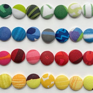 May include: A selection of fabric-covered buttons in a range of colours and patterns, such as green, blue, red, and yellow. The buttons are arranged in neat rows, displaying a variety of designs and textures. These buttons are suitable for sewing and craft projects.