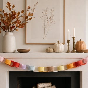 May include: A felt paper-chain style garland featuring interlocking loops in a gradient of colours including deep red, pink, orange, yellow, light blue, and tan. The garland is draped horizontally across the front of a fireplace mantel.