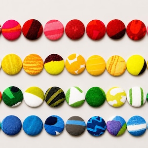 May include: A collection of thirty-six circular fabric-covered buttons arranged in four horizontal rows. The buttons feature a variety of colors including red, pink, yellow, green, and blue, with diverse patterns such as stripes, dots, and abstract shapes.