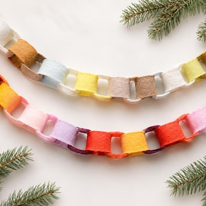 May include: Two decorative garlands made of interlocking felt loops in a variety of colours including red, orange, yellow, pink, purple, white, and tan. The chains are arranged in curved lines and feature a soft, textured material finish.