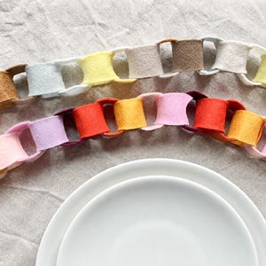May include: Two decorative felt paper chains arranged in a curved formation. The top chain features neutral tones of white, beige, tan, and pale yellow. The bottom chain displays vibrant colours including red, orange, yellow, pink, and purple.