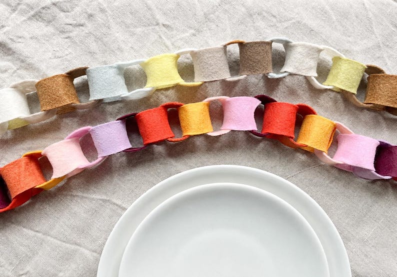 May include: Two decorative felt paper chains arranged in a curved formation. The top chain features neutral tones of white, beige, tan, and pale yellow. The bottom chain displays vibrant colours including red, orange, yellow, pink, and purple.