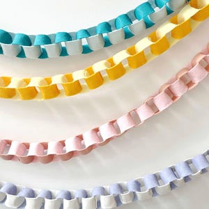 May include: Close-up of four colorful felt garlands. The garlands are in shades of teal, yellow, pink, and lavender. Each garland has a unique design with alternating felt balls and cut-out sections, creating a decorative and textured appearance.