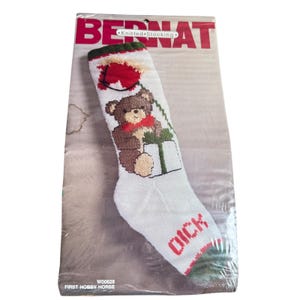 May include: A white Bernat knitted stocking in original packaging featuring a brown teddy bear holding a green gift with a red bow. The stocking has a green cuff and toe, with the name Dick embroidered in red near the bottom. The packaging includes the text Bernat, Knitted Stocking, and First Hobby Horse.