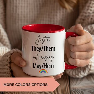 May include: A white ceramic mug with a red rim and handle. The mug has black text that reads "Just a They/Them out causing May/Hem" with a rainbow underneath.