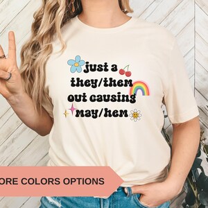 May include: Cream-colored t-shirt with the text "just a they/them out causing may/hem" in black, with colorful graphics including a blue flower, cherries, a rainbow, a star, and a daisy. The shirt has short sleeves.