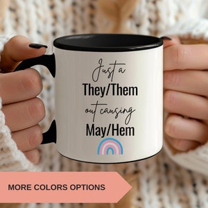 May include: White ceramic mug with a black rim and handle. The mug has black text that reads "Just a They/Them out causing May/Hem" with a rainbow in the colors of the transgender pride flag.