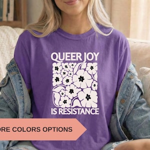 May include: A purple t-shirt with the text "QUEER JOY IS RESISTANCE" in white, along with a floral design. The shirt is part of a clothing collection and is available in more colors.