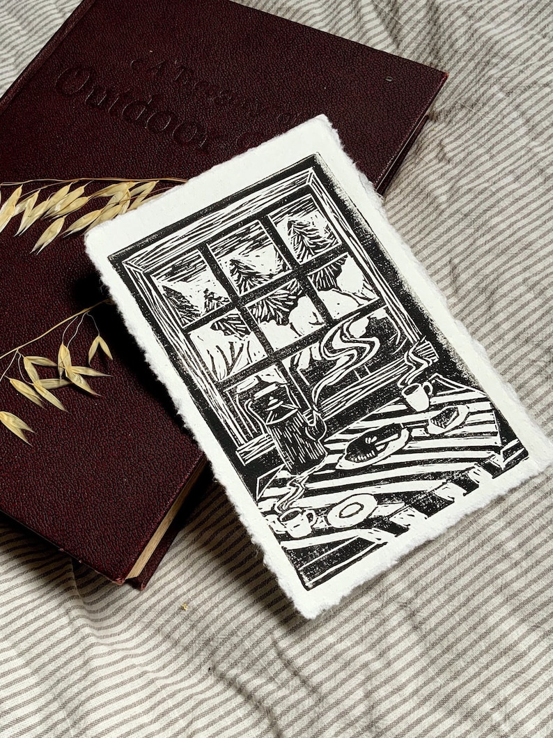 May include: Black and white linocut print of a window with a view of a snowy forest. The window is framed in wood and there is a table with a teapot, cups, and a plate in the foreground.