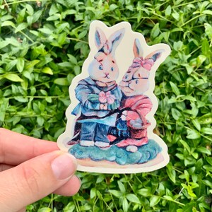 May include: A sticker featuring two cartoon rabbits. One rabbit wears a blue jacket and holds pink flowers. The other rabbit wears a pink dress and holds a basket. The sticker has a cream-colored background and is set against a green leafy background.