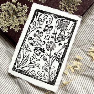 Pode incluir: Impress&atilde;o em lin&oacute;leo preto e branco de um desenho floral com v&aacute;rias flores e folhas. A impress&atilde;o &eacute; emoldurada com uma borda preta grossa.