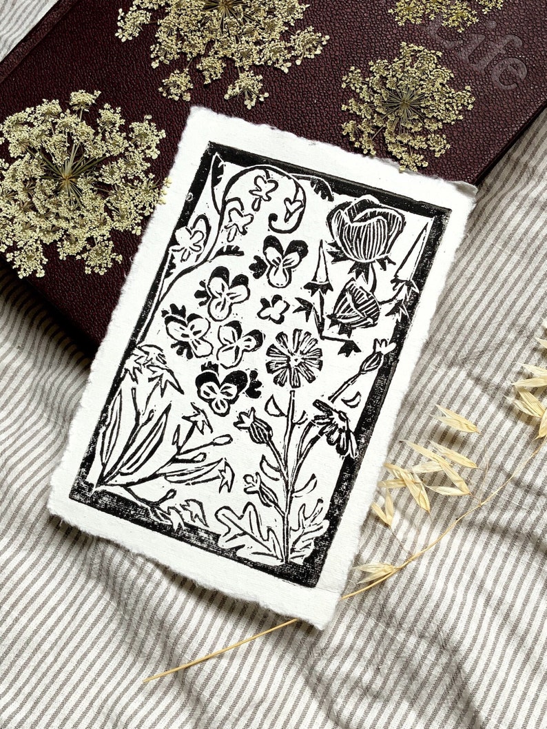 Pode incluir: Impress&atilde;o em lin&oacute;leo preto e branco de um desenho floral com v&aacute;rias flores e folhas. A impress&atilde;o &eacute; emoldurada com uma borda preta grossa.