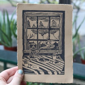 May include: A linocut print on brown paper depicting a window view of a snowy forest with a teapot, cups, and a plate of food on a table in the foreground.