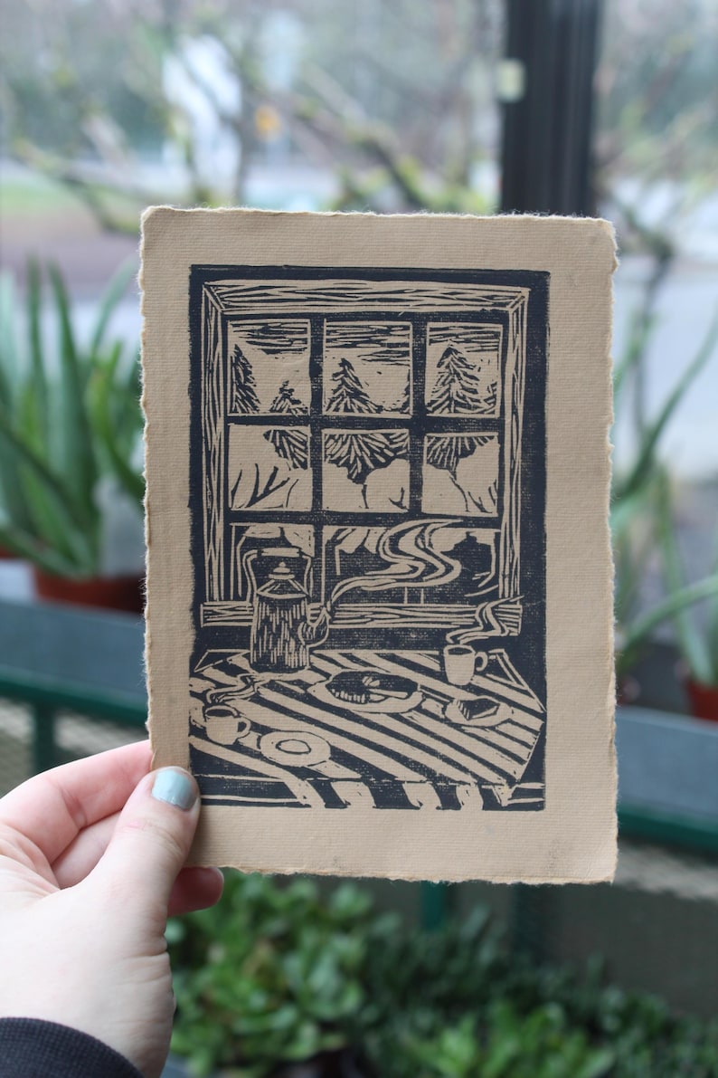 May include: A linocut print on brown paper depicting a window view of a snowy forest with a teapot, cups, and a plate of food on a table in the foreground.