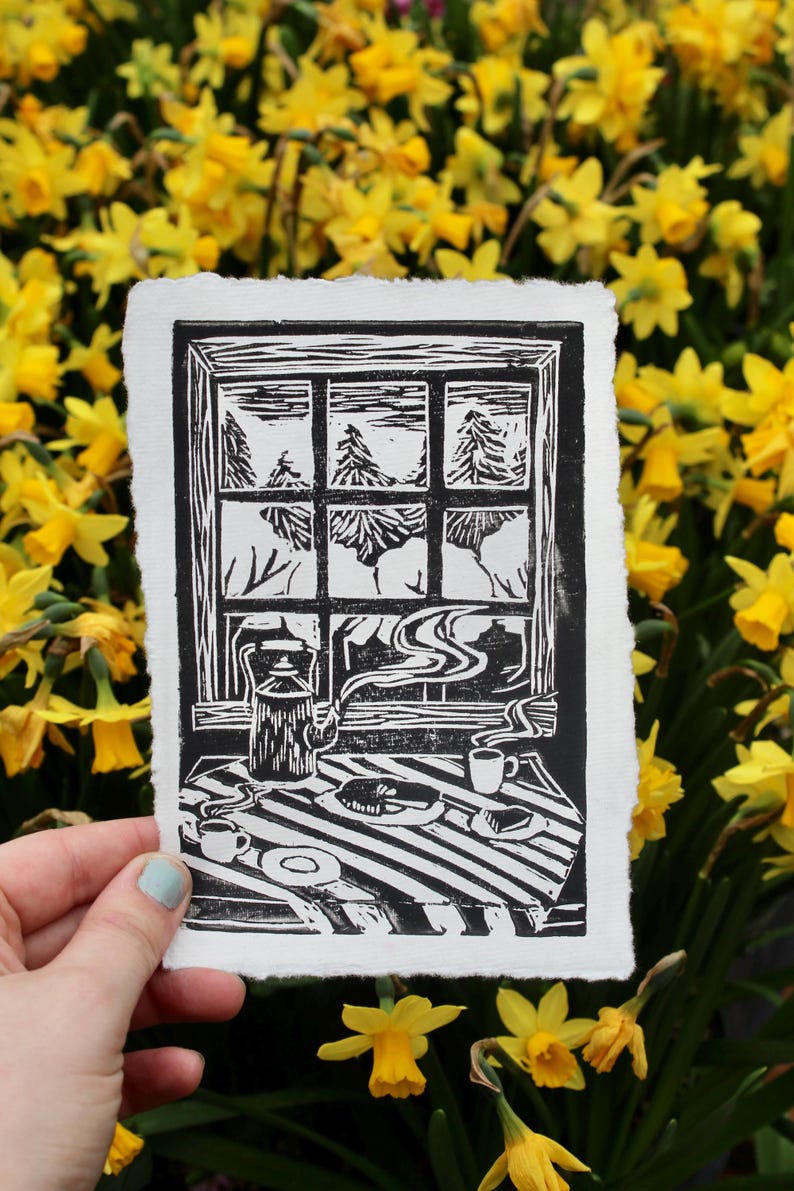 May include: Black and white linocut print of a window with a view of a snowy forest. A table with a teapot, cups, and a plate of food is in the foreground.