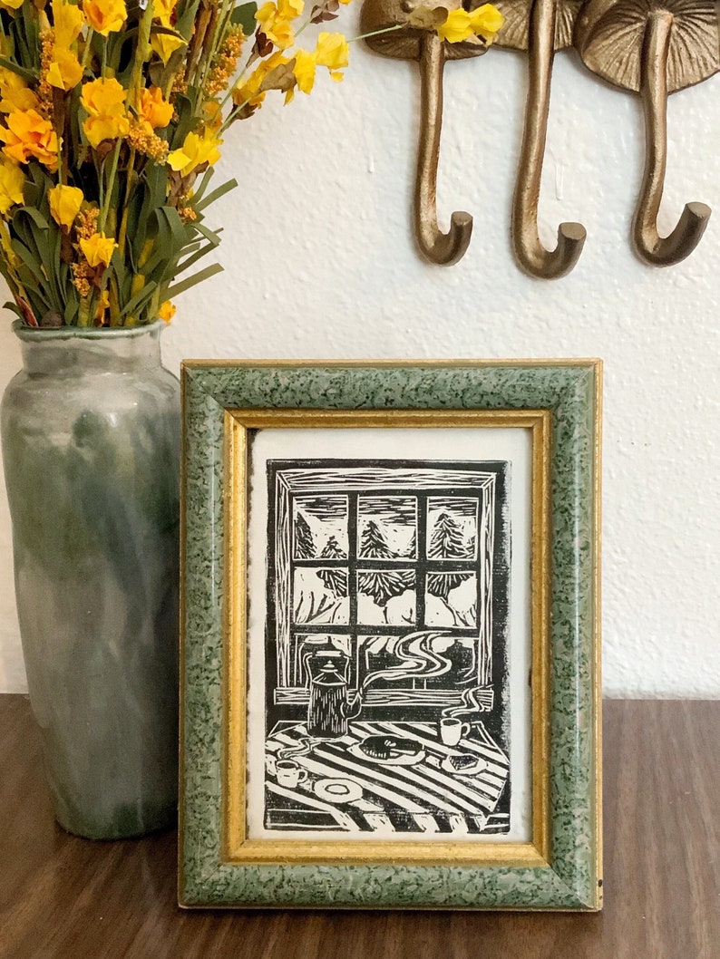 May include: A framed black and white woodblock print of a window scene with a teapot, cups, and a plate of food on a striped tablecloth. The window shows a snowy forest scene.