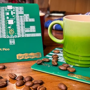 Könnte beinhalten: Ein grüner Raspberry Pi Pico Pinout-Diagramm auf einem Untersetzer mit dem Text "GurgieApps.com". Eine grüne Tasse mit einem hellgrünen Oberteil steht auf einem passenden Untersetzer mit Kaffeekörnern, die herumliegen.
