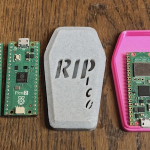 Puede incluir: Dos placas de circuito Pico 2 verdes y una carcasa gris en forma de tumba con "RIP" recortado. Una carcasa rosa en forma de ataúd contiene una placa de circuito verde. Sobre una superficie de madera.