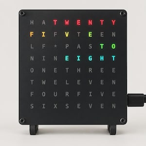 May include: A square black word clock with a grid of letters. The illuminated words "TWENTY", "FIFTEEN", "PAST", "TO", and "EIGHT" are displayed in red, yellow, green, and blue, indicating the time. The clock has a power cord attached to the side.