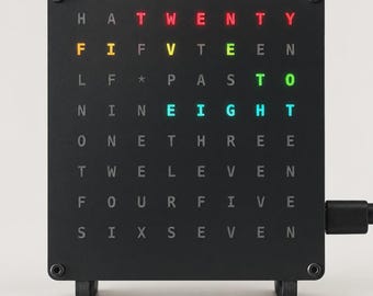 Pode incluir: Um relógio de palavras quadrado preto com uma grade de letras. As palavras iluminadas "TWENTY", "FIFTEEN", "PAST", "TO" e "EIGHT" aparecem em vermelho, amarelo, verde e azul, indicando a hora. O relógio tem um cabo de energia lateral.