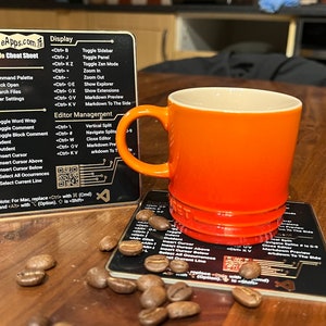 May include: A black and gold VS Code cheat sheet with a QR code and a mug of orange and yellow coffee on a wooden table with coffee beans scattered around.