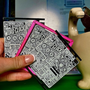 Può includere: Scheda a circuito Word Clock Matrix in bianco e nero con cornice rosa. Il design presenta le parole "WORD-CLOCK" e "MATRIX" in uno stile tecnico e geometrico. Il prodotto è tenuto in mano.