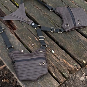 Puede incluir: Bolso de pecho marrón con múltiples bolsillos y un diseño único. El bolso presenta líneas horizontales, una cremallera negra y correas negras ajustables con herrajes metálicos dorados. Diseñado para llevarse sobre el pecho.