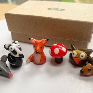 May include: A set of six miniature woodland animal figurines made of polymer clay. The set includes a fox, a badger, a mouse, an owl, a mushroom, and a hedgehog.