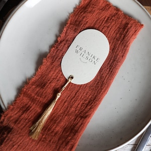 May include: A table setting with a white plate, a burnt orange linen napkin, and a name card. The oval name card has the text "FRANKE WILSON WELCOME" and a gold tassel. Silver forks are on the left side of the plate.