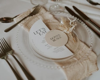 May include: A white table setting with a beige napkin, a clear beaded charger plate, and a white menu card with the text "Let's Eat". A small round place card with the text "Donya Beale" is on top of the napkin.