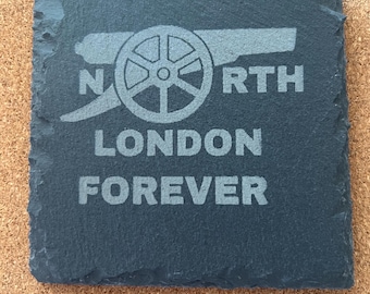 May include: A square, dark gray slate coaster with the words "NORTH LONDON FOREVER" etched in a light gray font. The design includes a cannon graphic, a symbol of the Arsenal Football Club.